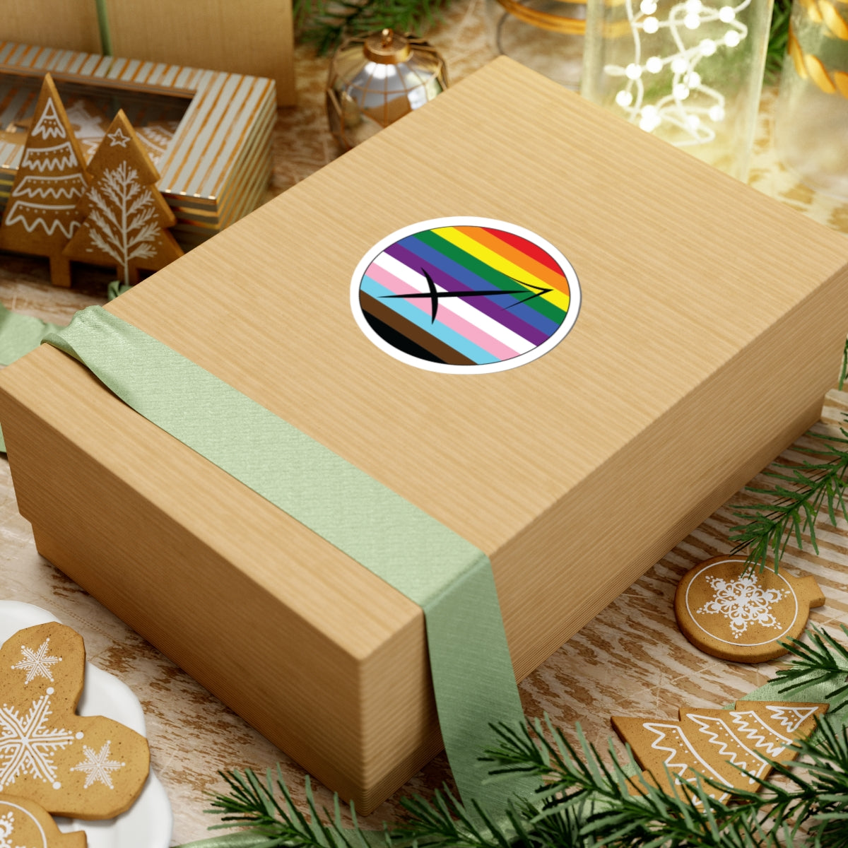 Gift box with a green ribbon on a festive background with cookies and Christmas decorations. Sagittarius Progress pride sticker centered on the box.