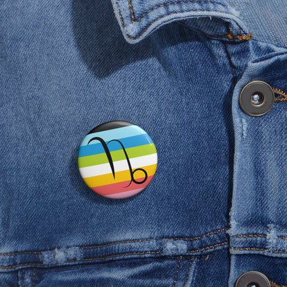 Close-up of a white circular pride button featuring the Capricorn zodiac glyph centered over a queer pride flag background in pinks, yellow, white, green, blues, and black, attached to a blue denim jacket at the right chest with the button clearly visible.