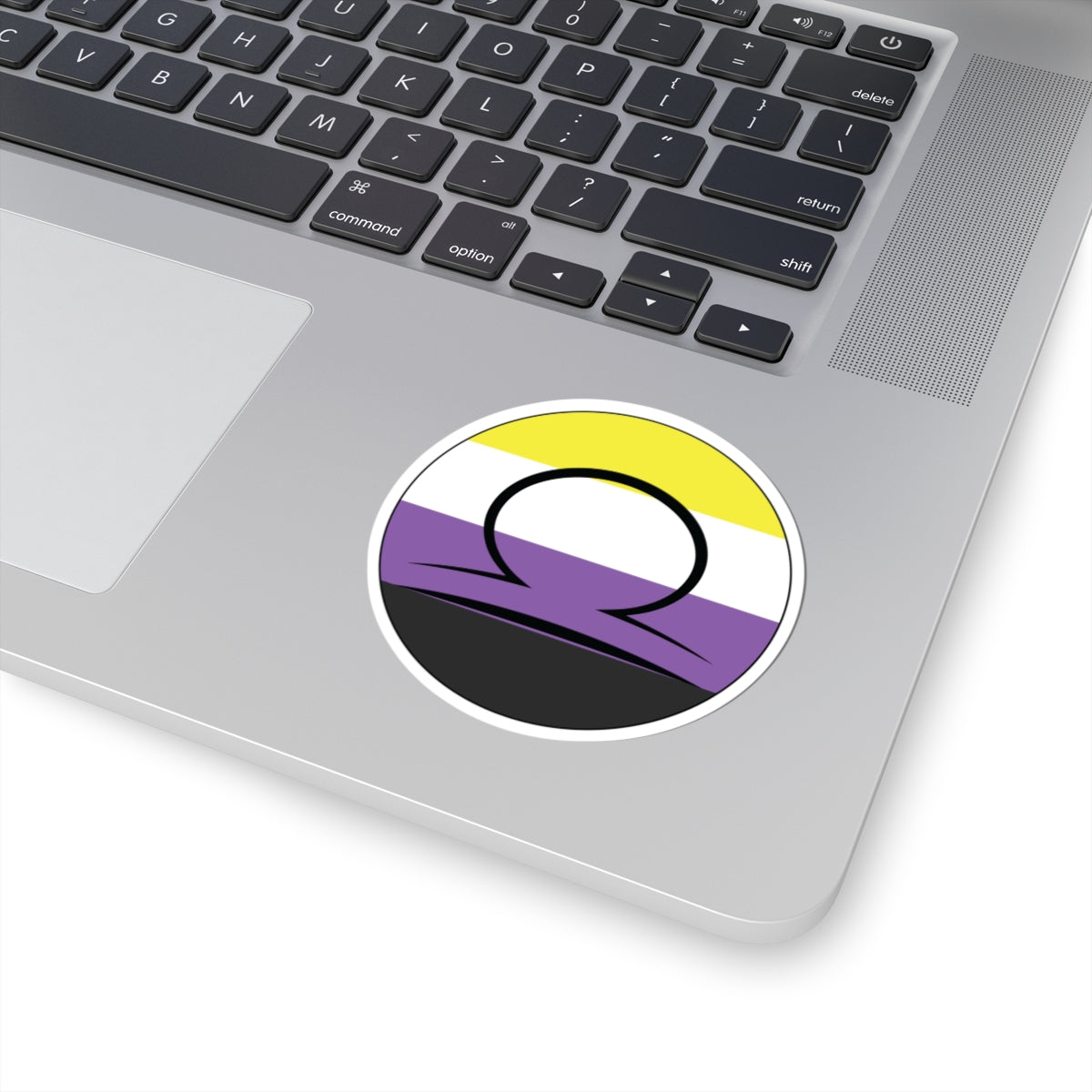 Close-up of the bottom right corner of a silver laptop featuring a 3 inch by 3 inch circular zodiac sticker with a centered Libra glyph on a Nonbinary pride flag style background applied to the surface.