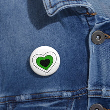 Close-up of a white circular pride button featuring a nested heart design inspired by the Neutrois pride flag in white, green and black, attached to a blue denim jacket at the right chest with the button clearly visible.
