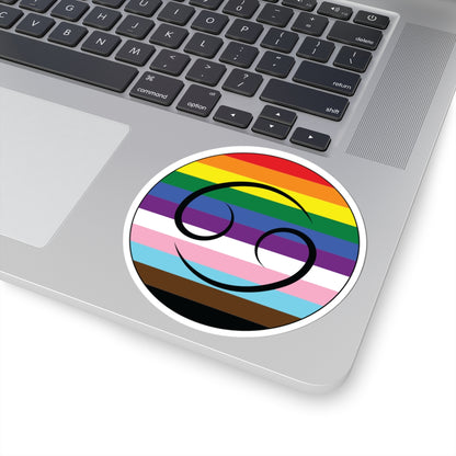 Close-up of the bottom right corner of a silver laptop featuring a 4 inch by 4 inch circular zodiac sticker with a centered Cancer glyph on a progress pride flag style background applied to the surface.