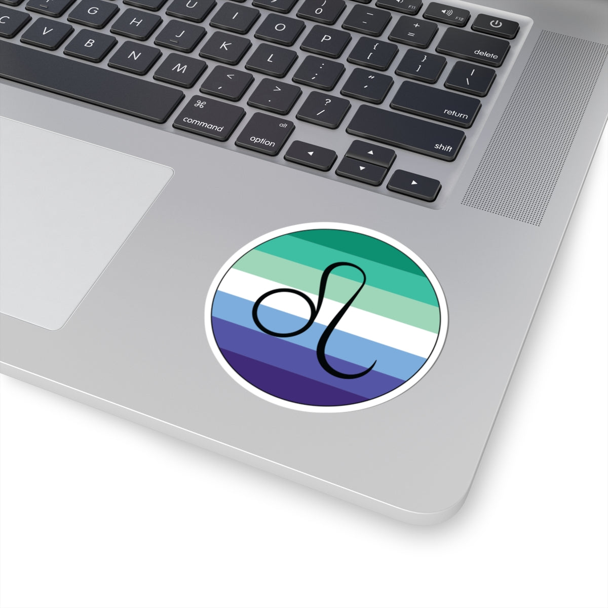Close-up of the bottom right corner of a silver laptop featuring a 3 inch by 3 inch circular zodiac sticker with a centered Leo glyph on a Gay Men pride flag style background applied to the surface.