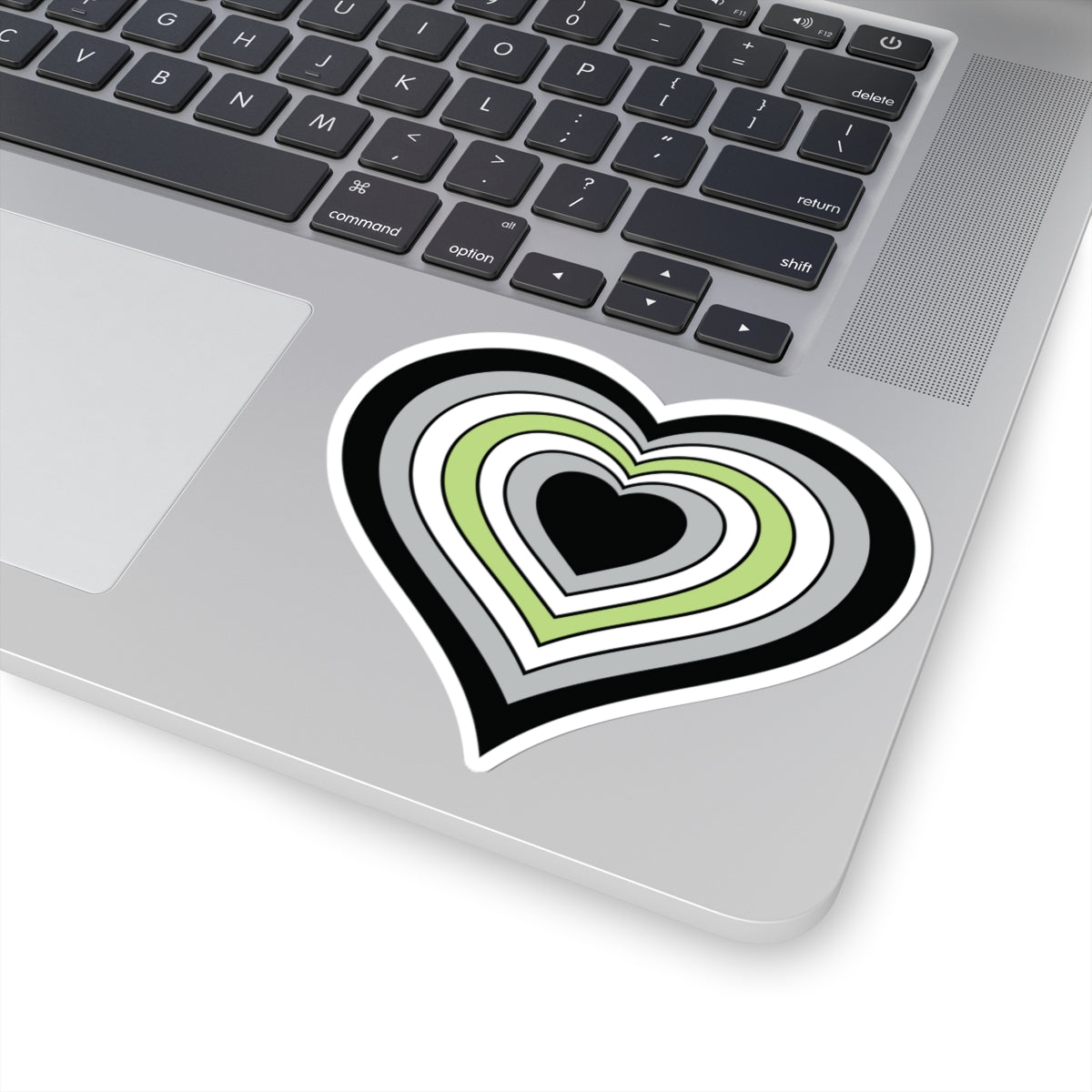 Close-up of the bottom right corner of a silver laptop featuring a 4 inch by 4 inch pride flag heart sticker in an Agender flag style applied to the surface.