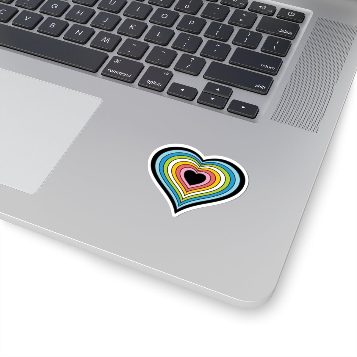 Close-up of the bottom right corner of a silver laptop featuring a 2 inch by 2 inch pride flag heart sticker in a Queer flag style applied to the surface.