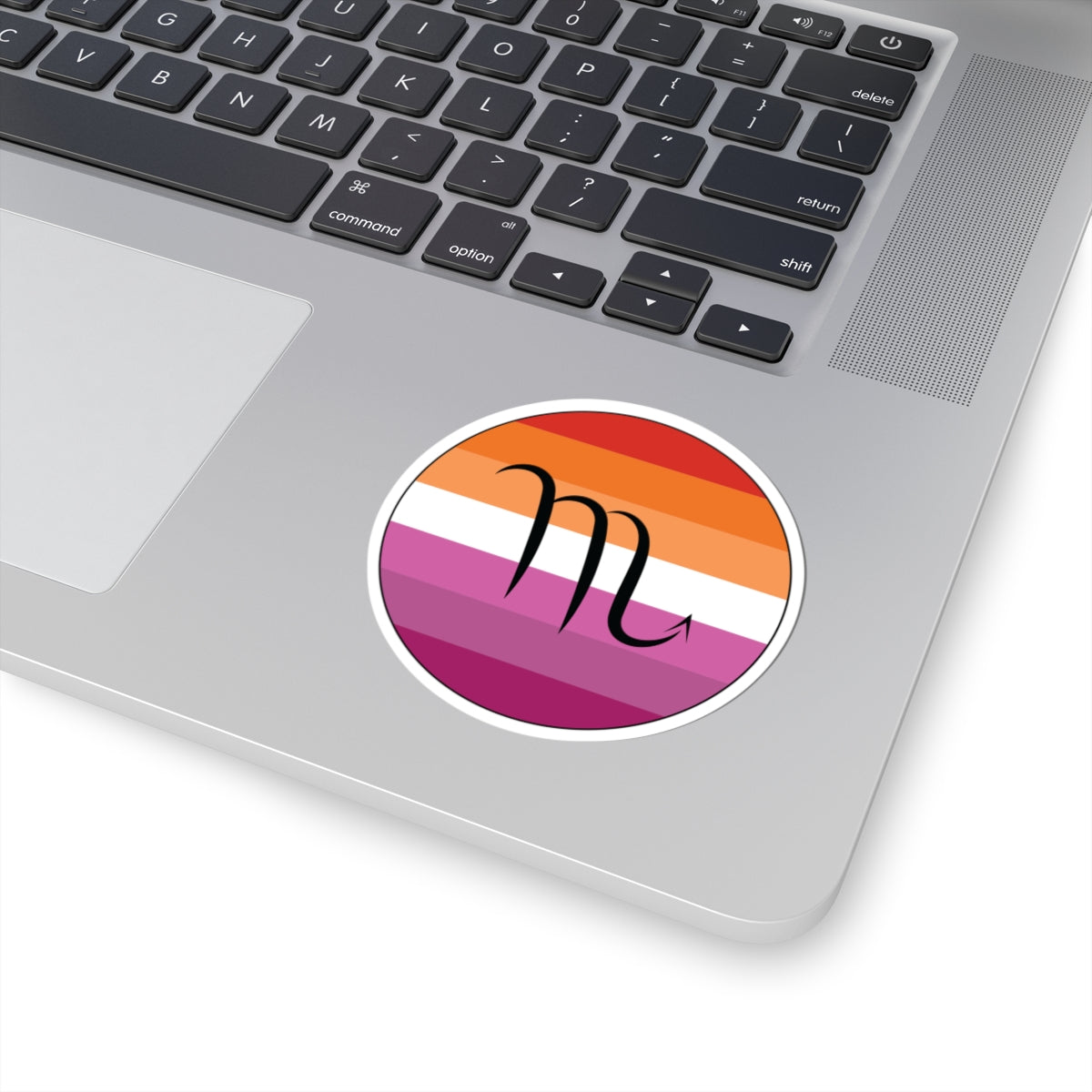 Close-up of the bottom right corner of a silver laptop featuring a 3 inch by 3 inch circular zodiac sticker with a centered Scorpio glyph on a Lesbian pride flag style background applied to the surface.
