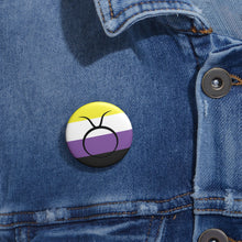 Close-up of a white circular pride button featuring the Taurus zodiac glyph centered over a nonbinary pride flag background in yellow, white, purple, and black, attached to a blue denim jacket at the right chest with the button clearly visible.