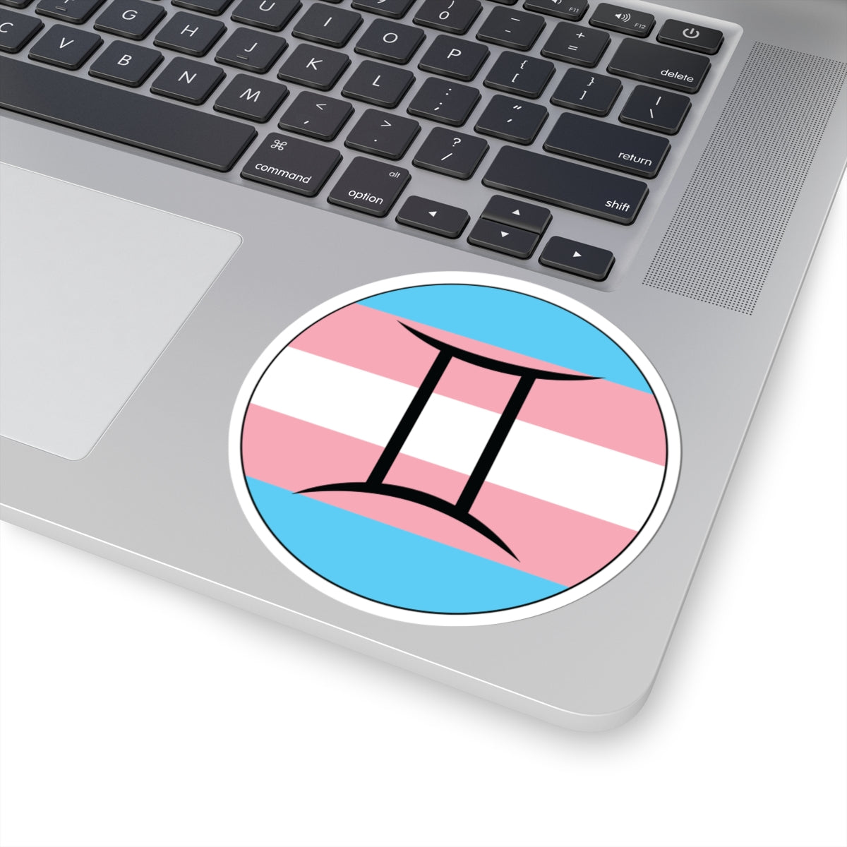 Close-up of the bottom right corner of a silver laptop featuring a 4 inch by 4 inch circular zodiac sticker with a centered Gemini glyph on a Transgender pride flag style background applied to the surface.
