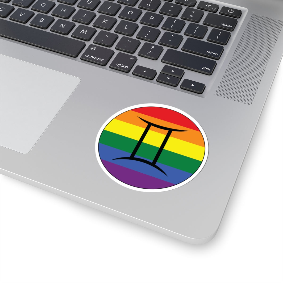 Close-up of the bottom right corner of a silver laptop featuring a 3 inch by 3 inch circular zodiac sticker with a centered Gemini glyph on a Rainbow pride flag style background applied to the surface.