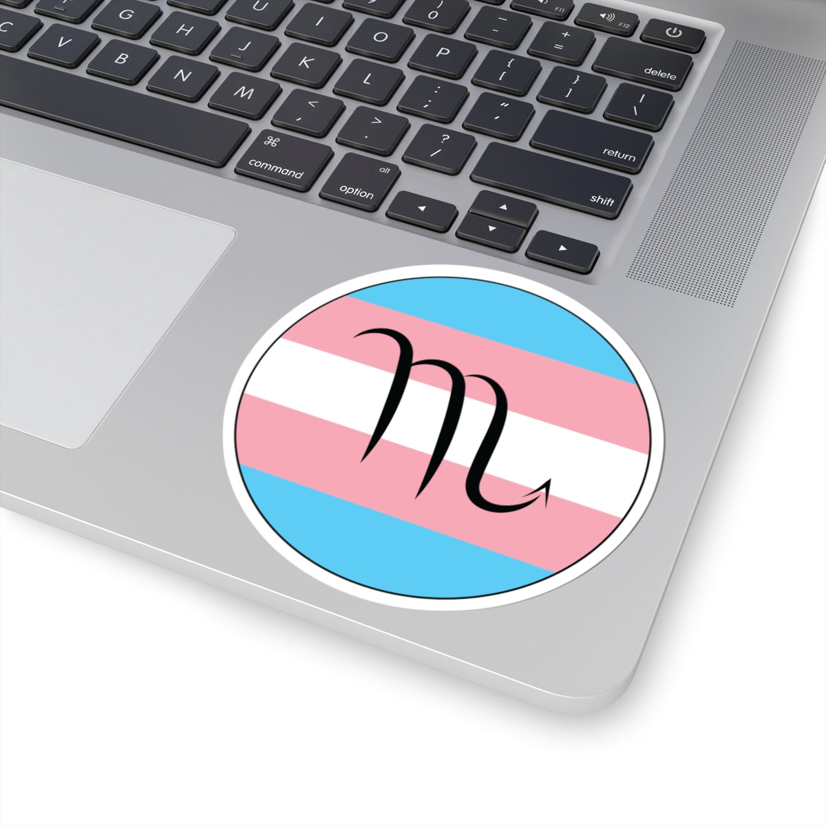 Close-up of the bottom right corner of a silver laptop featuring a 4 inch by 4 inch circular zodiac sticker with a centered Scorpio glyph on a Transgender pride flag style background applied to the surface.