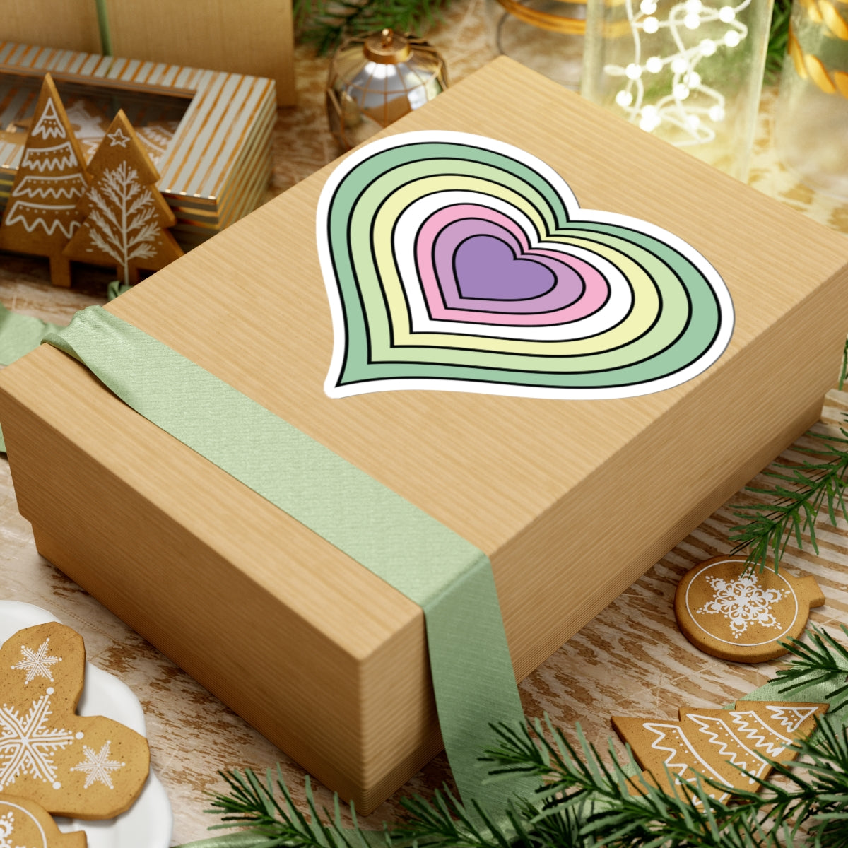 Gift box with a green ribbon on a festive background with cookies and Christmas decorations. Heart-shaped Genderfae pride sticker centered on the box.