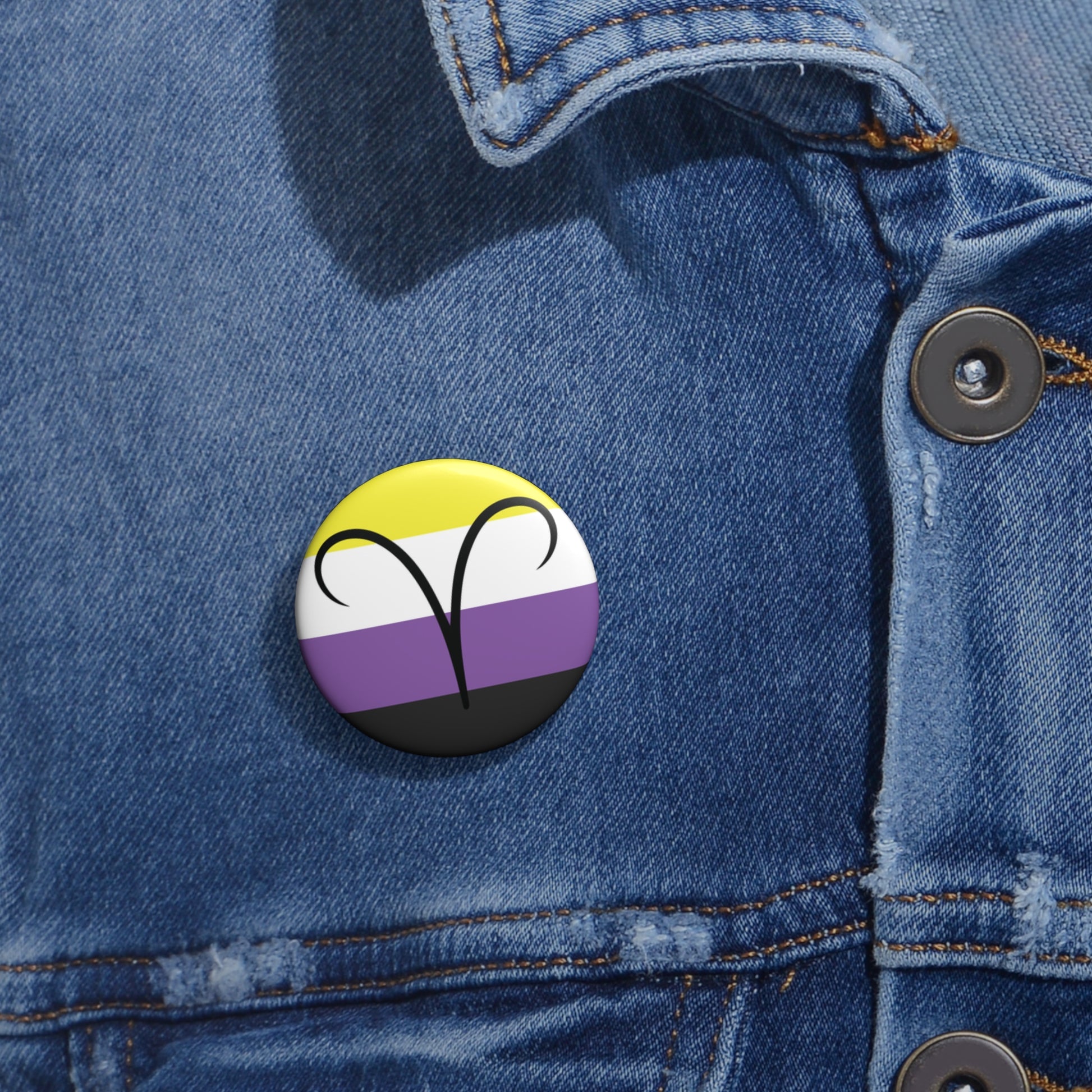 Close-up of a white circular pride button featuring the Aires zodiac glyph centered over a nonbinary pride flag background in yellow, white, purple, and black, attached to a blue denim jacket at the right chest with the button clearly visible.