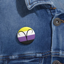 Close-up of a white circular pride button featuring the Aires zodiac glyph centered over a nonbinary pride flag background in yellow, white, purple, and black, attached to a blue denim jacket at the right chest with the button clearly visible.