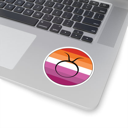 Close-up of the bottom right corner of a silver laptop featuring a 3 inch by 3 inch circular zodiac sticker with a centered Taurus glyph on a Lesbian pride flag style background applied to the surface.