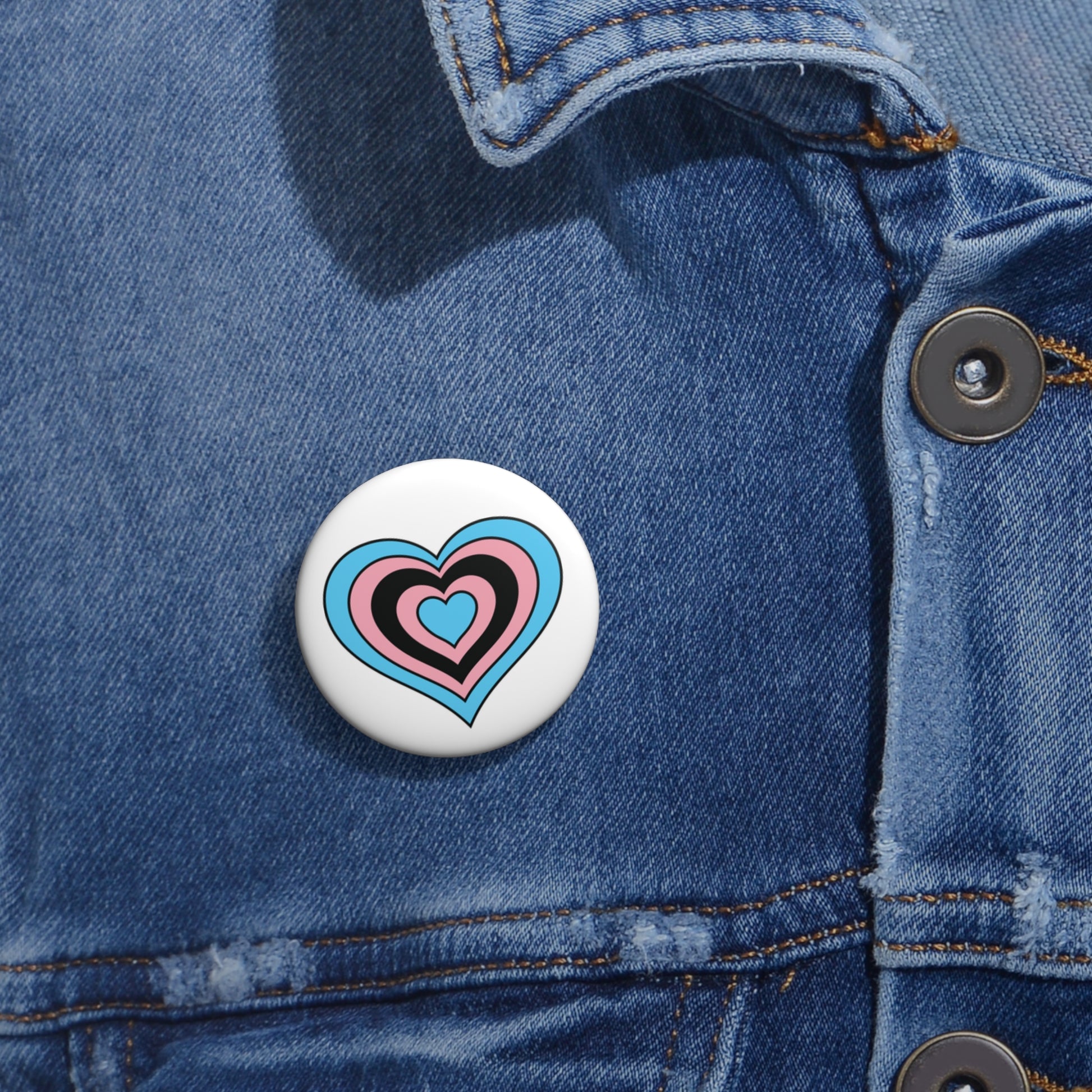 Close-up of a white circular pride button featuring a nested heart design inspired by the Black Trans pride flag in blue, pink and black, attached to a blue denim jacket at the right chest with the button clearly visible.