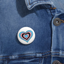 Close-up of a white circular pride button featuring a nested heart design inspired by the Black Trans pride flag in blue, pink and black, attached to a blue denim jacket at the right chest with the button clearly visible.