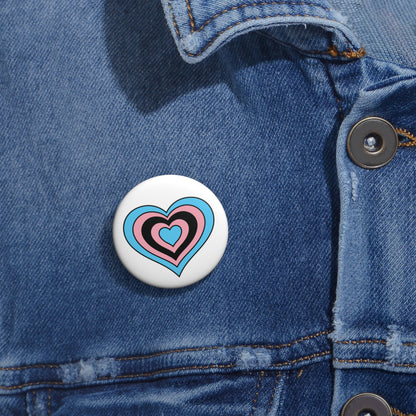 Close-up of a white circular pride button featuring a nested heart design inspired by the Black Trans pride flag in blue, pink and black, attached to a blue denim jacket at the right chest with the button clearly visible.