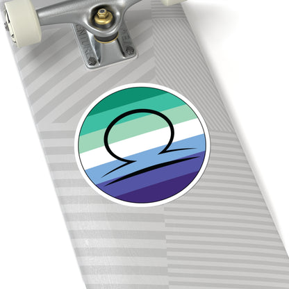 Close-up of the bottom right corner of a silver laptop featuring a 6 inch by 6 inch circular zodiac sticker with a centered Libra glyph on a Gay Men pride flag style background applied to the surface.