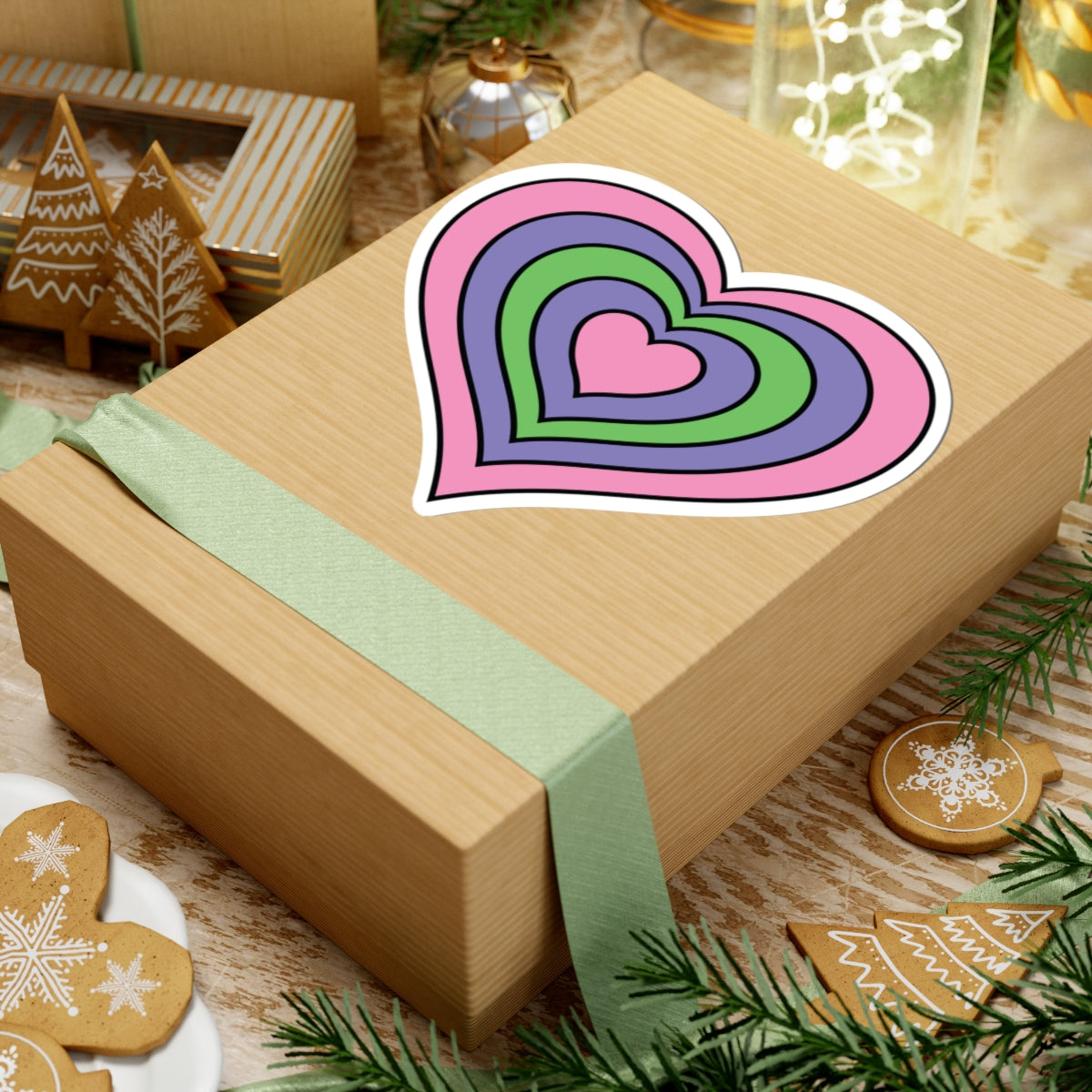 Gift box with a green ribbon on a festive background with cookies and Christmas decorations. Heart-shaped Trigender pride sticker centered on the box.