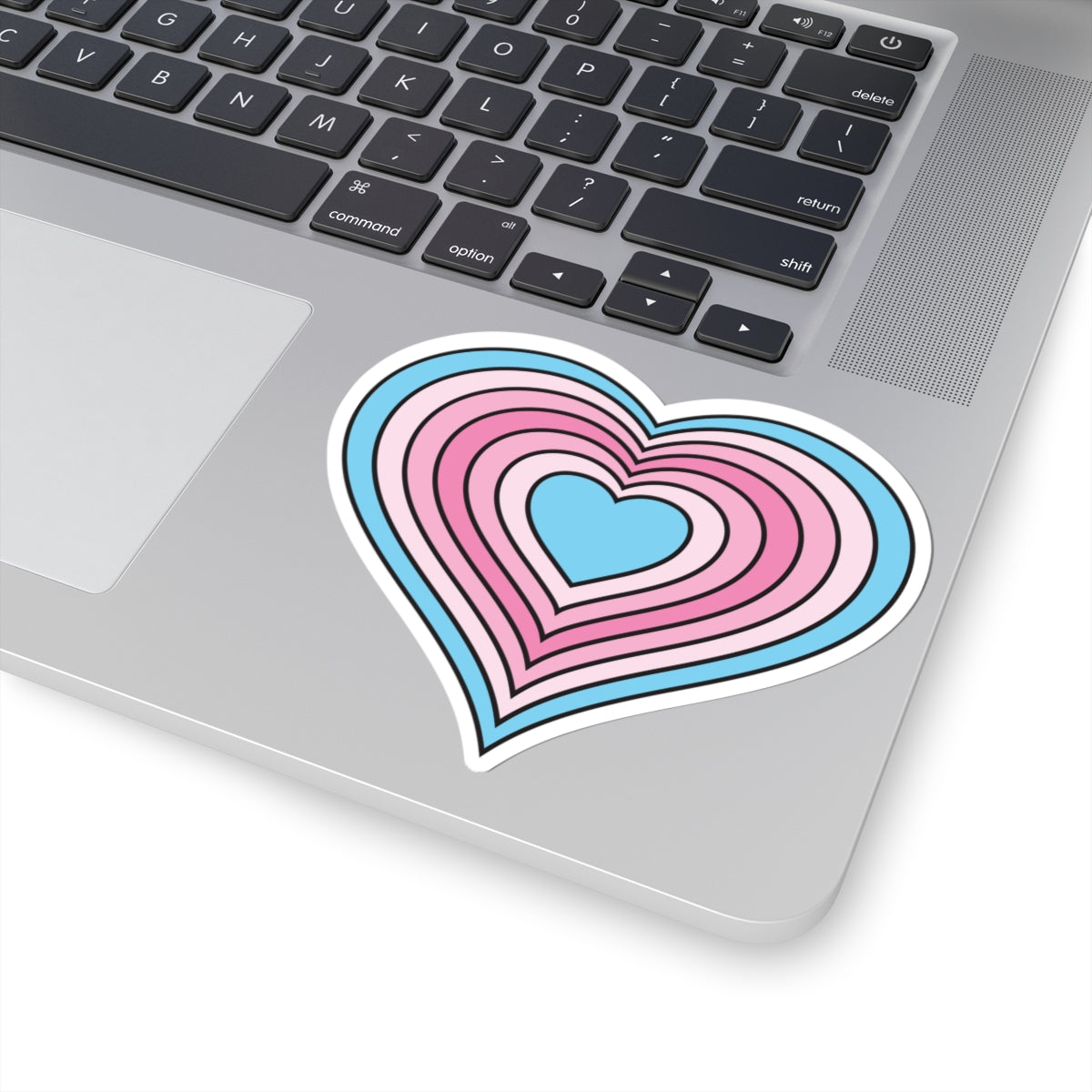 Close-up of the bottom right corner of a silver laptop featuring a 4 inch by 4 inch pride flag heart sticker in a Transfem flag style applied to the surface.