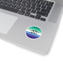Close-up of the bottom right corner of a silver laptop featuring a 2 inch by 2 inch circular zodiac sticker with a centered Aquarius glyph on a Gay Men pride flag style background applied to the surface.