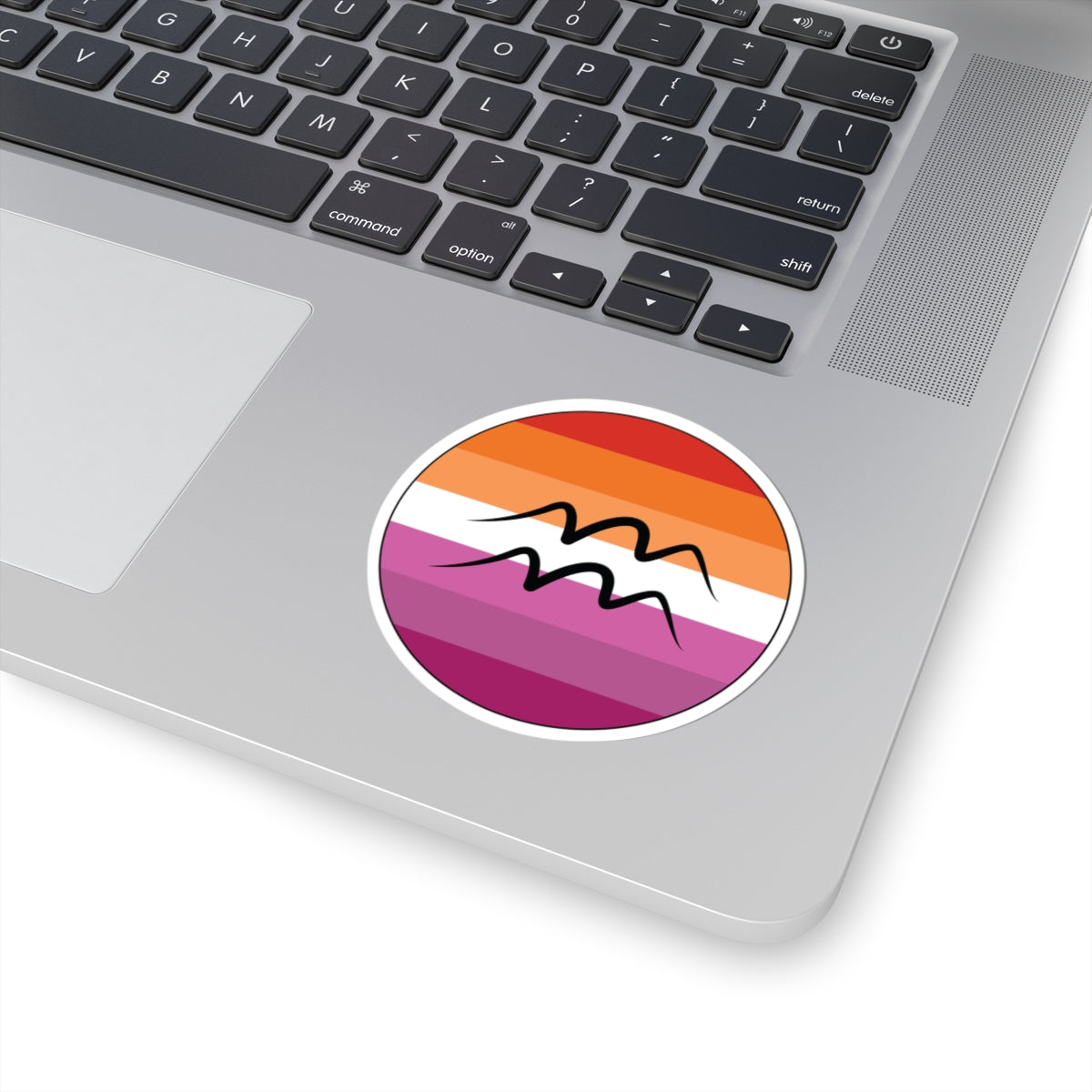 Close-up of the bottom right corner of a silver laptop featuring a 3 inch by 3 inch circular zodiac sticker with a centered Aquarius glyph on a Lesbian pride flag style background applied to the surface.