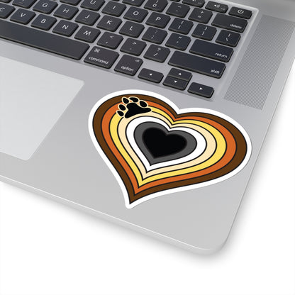 Close-up of the bottom right corner of a silver laptop featuring a 4 inch by 4 inch pride flag heart sticker in a Bear pride flag style applied to the surface.