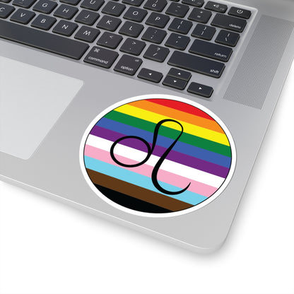 Close-up of the bottom right corner of a silver laptop featuring a 4 inch by 4 inch circular zodiac sticker with a centered Leo glyph on a progress pride flag style background applied to the surface.