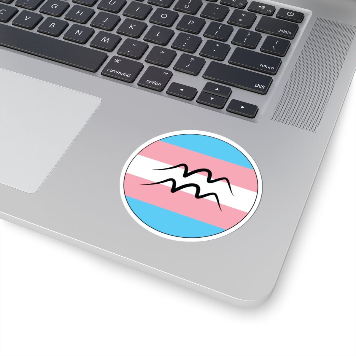 Close-up of the bottom right corner of a silver laptop featuring a 3 inch by 3 inch circular zodiac sticker with a centered Aquarius glyph on a Transgender pride flag style background applied to the surface.