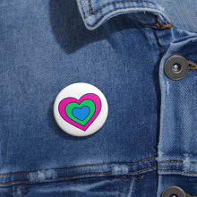 Close-up of a white circular pride button featuring a nested heart design inspired by the Polysexual pride flag in pink, green and blue, attached to a blue denim jacket at the right chest with the button clearly visible.
