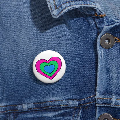 Close-up of a white circular pride button featuring a nested heart design inspired by the Polysexual pride flag in pink, green and blue, attached to a blue denim jacket at the right chest with the button clearly visible.