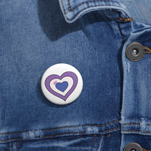 Close-up of a white circular pride button featuring a nested heart design inspired by the Drag pride flag in lavender, white, and blue with a pink crown, attached to a blue denim jacket at the right chest with the button clearly visible.