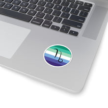Close-up of the bottom right corner of a silver laptop featuring a 2 inch by 2 inch circular zodiac sticker with a centered Capricorn glyph on a Gay Men pride flag style background applied to the surface.