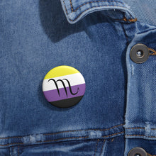 Close-up of a white circular pride button featuring the Scorpio zodiac glyph centered over a nonbinary pride flag background in yellow, white, purple, and black, attached to a blue denim jacket at the right chest with the button clearly visible.