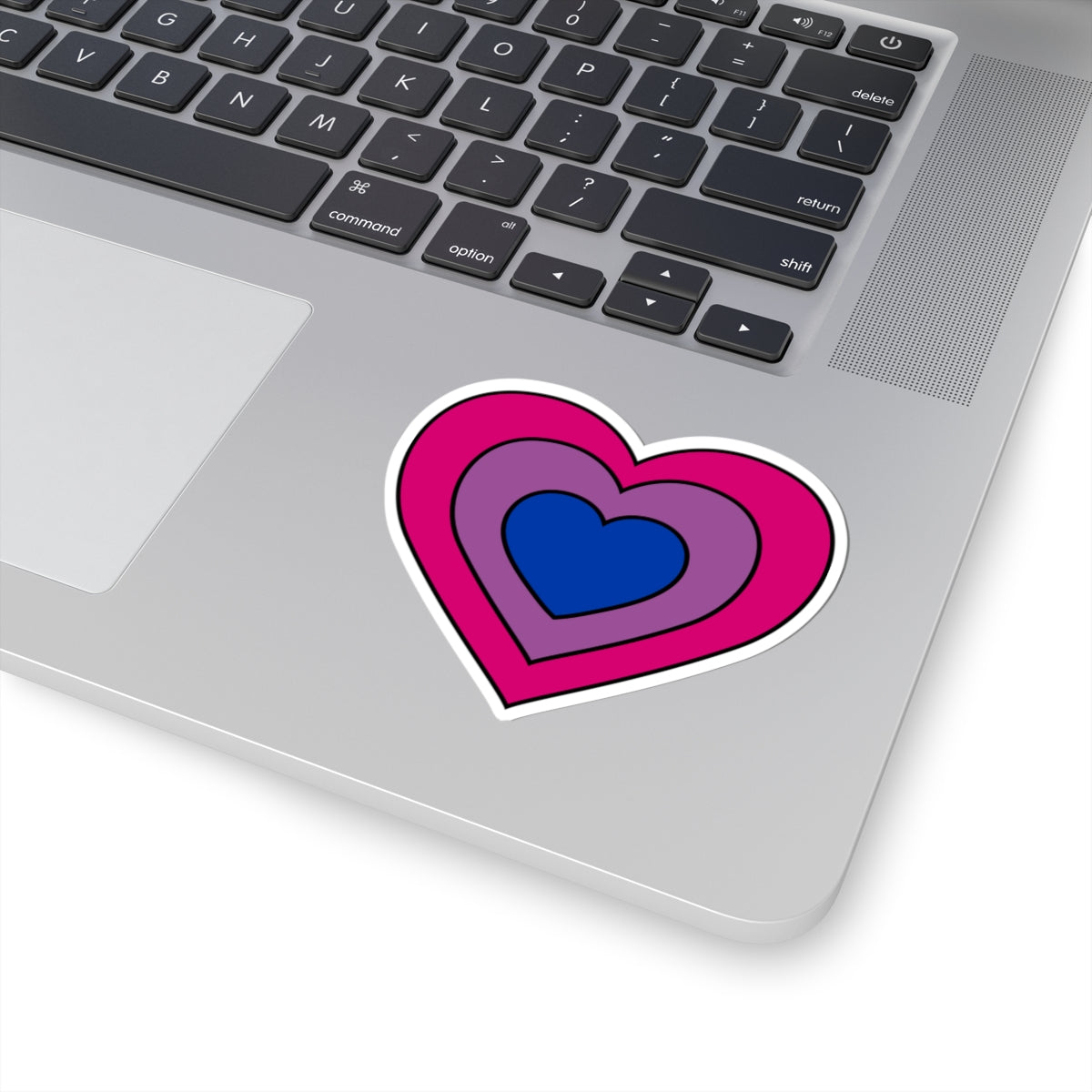 Close-up of the bottom right corner of a silver laptop featuring a 3 inch by 3 inch pride flag heart sticker in a Bisexual flag style applied to the surface.