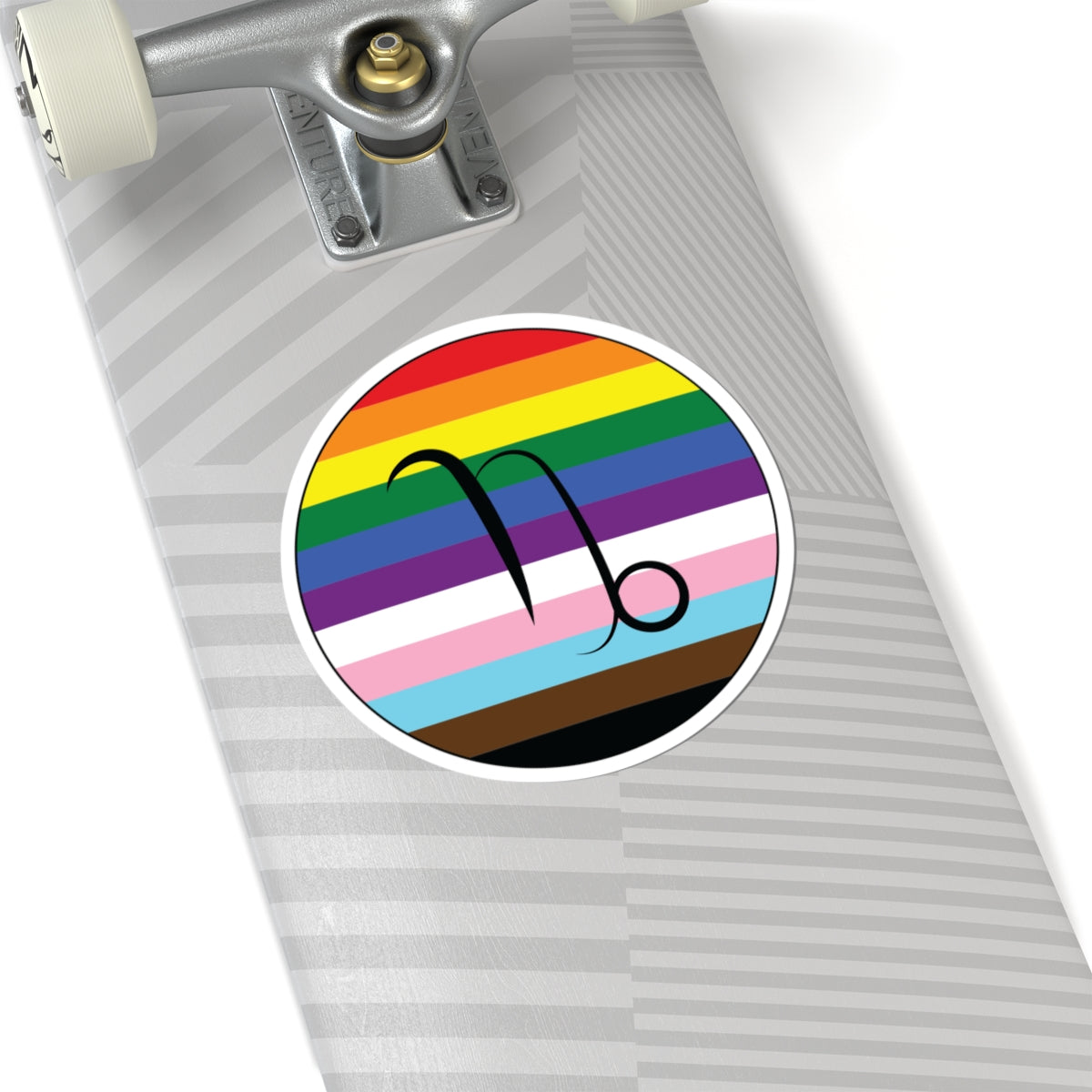 Close-up of the bottom of a silver skateboard featuring a 6 inch by 6 inch circular zodiac sticker with a centered Capricorn glyph on a progress pride flag style background applied to the surface.