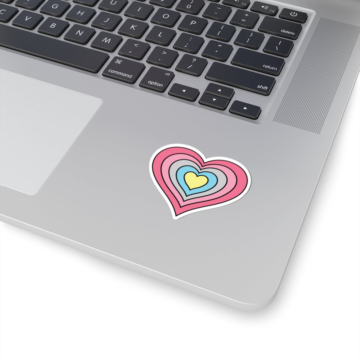Close-up of the bottom right corner of a silver laptop featuring a 2 inch by 2 inch pride flag heart sticker in a Genderflux flag style applied to the surface.