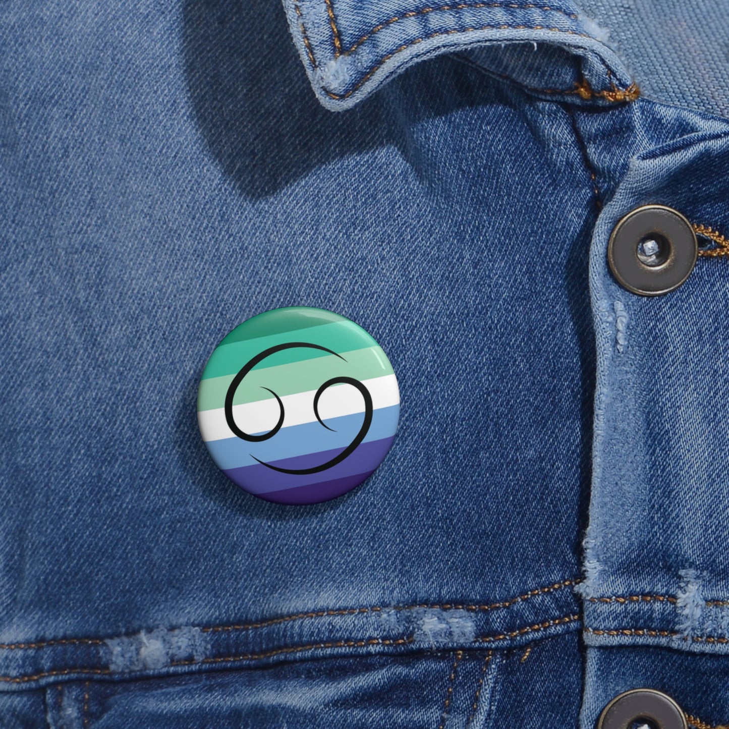 Close-up of a white circular pride button featuring the Cancer zodiac glyph centered over a gay men (MLM) pride flag background in greens/teals, white, and blues, attached to a blue denim jacket at the right chest with the button clearly visible.