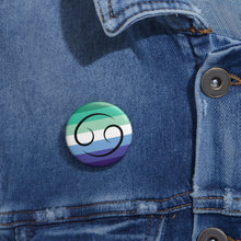 Close-up of a white circular pride button featuring the Cancer zodiac glyph centered over a gay men (MLM) pride flag background in greens/teals, white, and blues, attached to a blue denim jacket at the right chest with the button clearly visible.