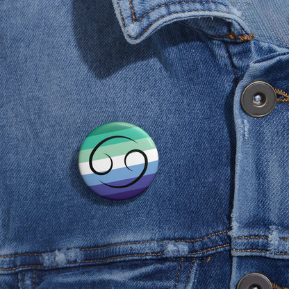 Close-up of a white circular pride button featuring the Cancer zodiac glyph centered over a gay men (MLM) pride flag background in greens/teals, white, and blues, attached to a blue denim jacket at the right chest with the button clearly visible.