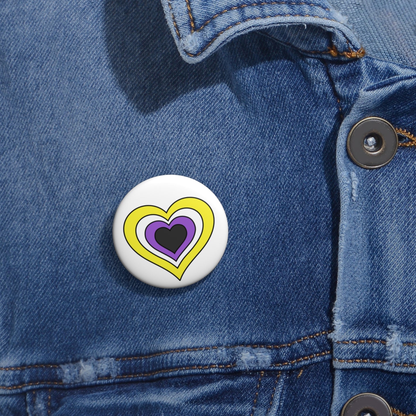 Close-up of a white circular pride button featuring a nested heart design inspired by the Nonbinary pride flag in yellow, white, purple and dark gray, attached to a blue denim jacket at the right chest with the button clearly visible.