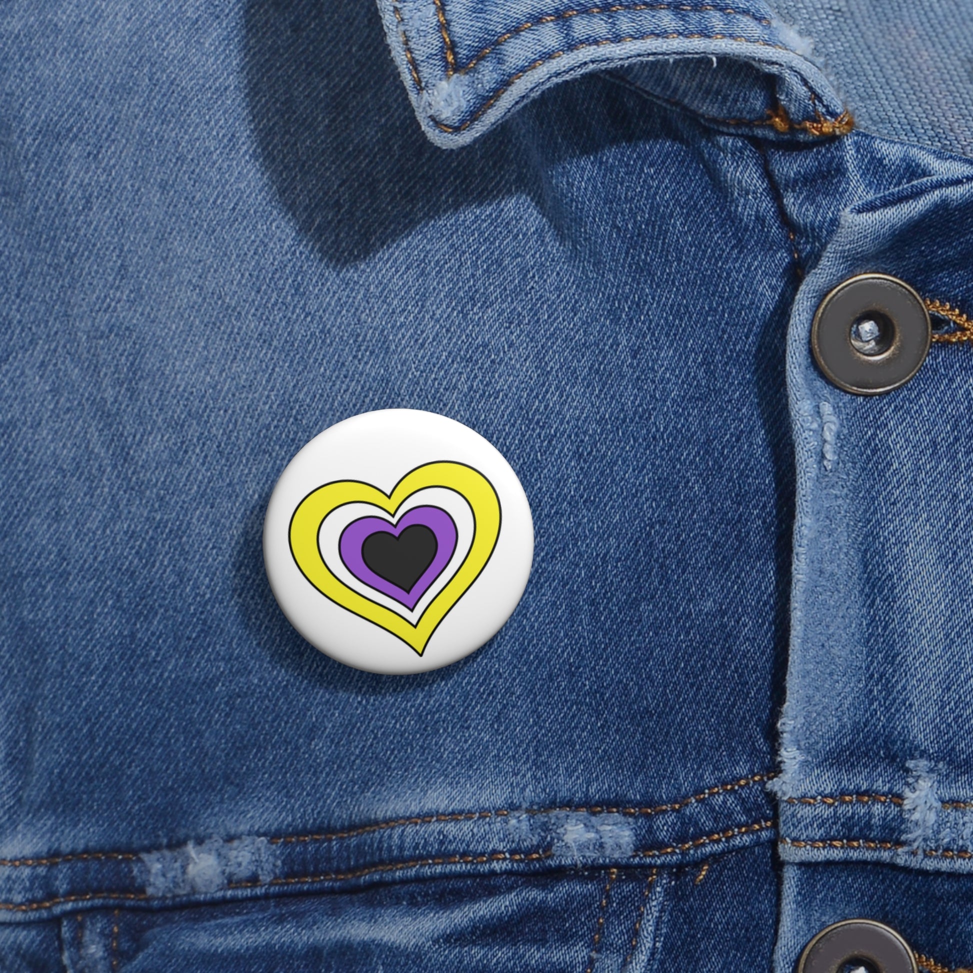 Close-up of a white circular pride button featuring a nested heart design inspired by the Nonbinary pride flag in yellow, white, purple and dark gray, attached to a blue denim jacket at the right chest with the button clearly visible.