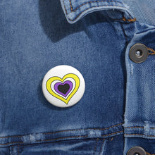 Close-up of a white circular pride button featuring a nested heart design inspired by the Nonbinary pride flag in yellow, white, purple and dark gray, attached to a blue denim jacket at the right chest with the button clearly visible.
