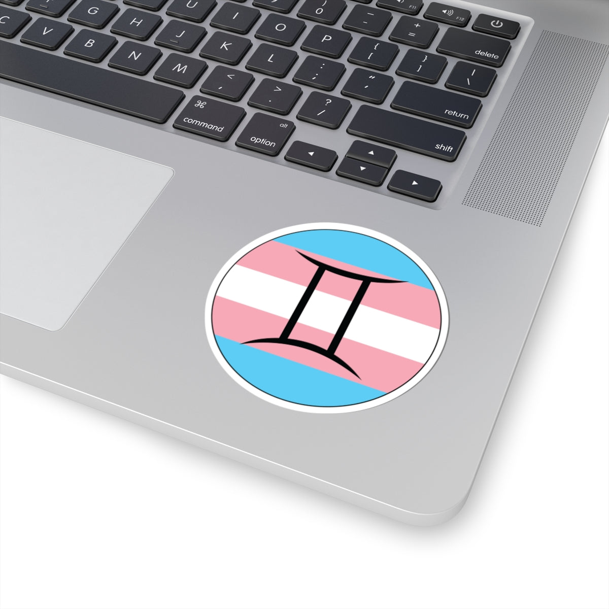 Close-up of the bottom right corner of a silver laptop featuring a 3 inch by 3 inch circular zodiac sticker with a centered Gemini glyph on a Transgender pride flag style background applied to the surface.