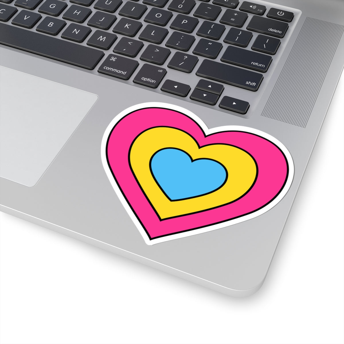 Close-up of the bottom right corner of a silver laptop featuring a 4 inch by 4 inch pride flag heart sticker in a Pansexual flag style applied to the surface.