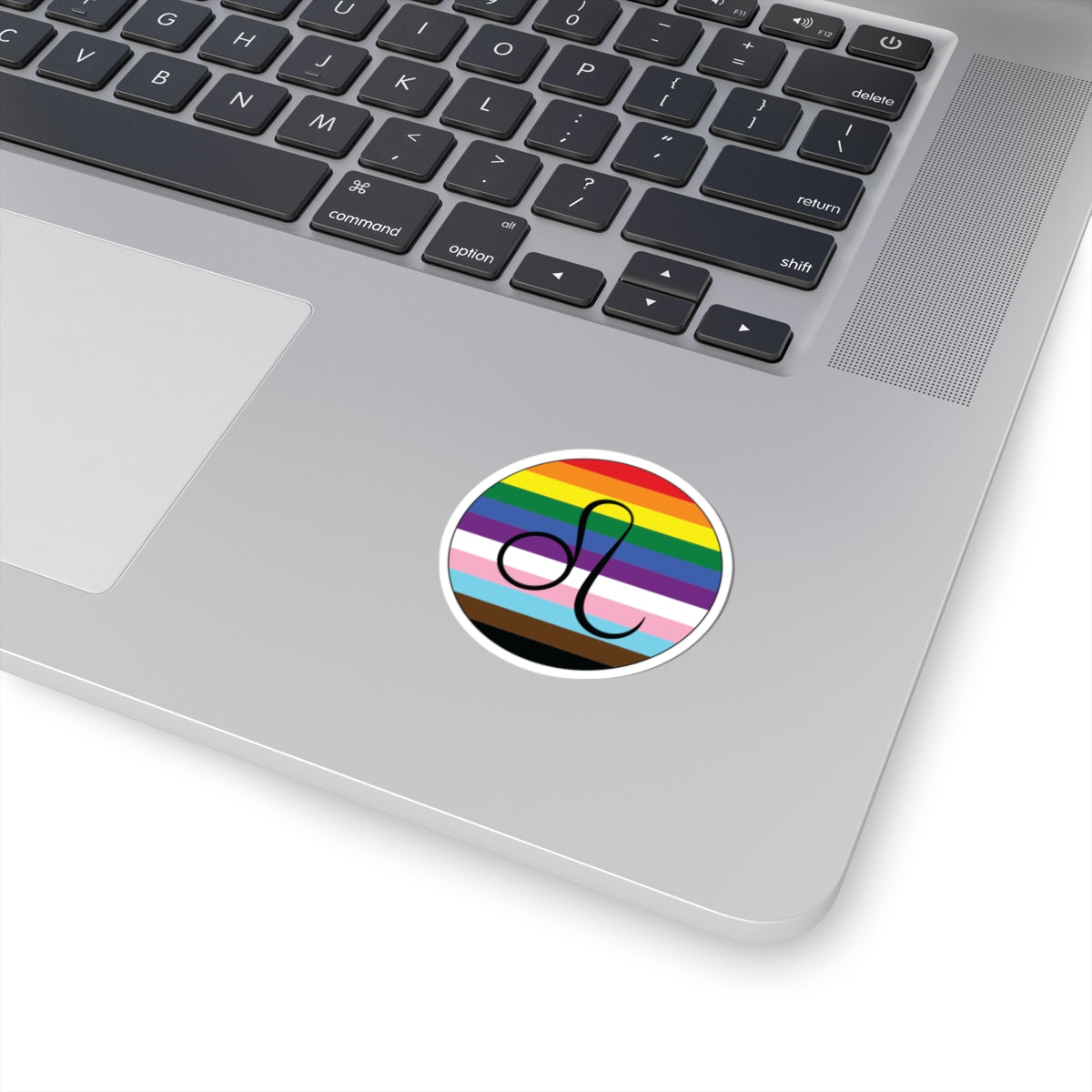 Close-up of the bottom right corner of a silver laptop featuring a 2 inch by 2 inch circular zodiac sticker with a centered Leo glyph on a progress pride flag style background applied to the surface.