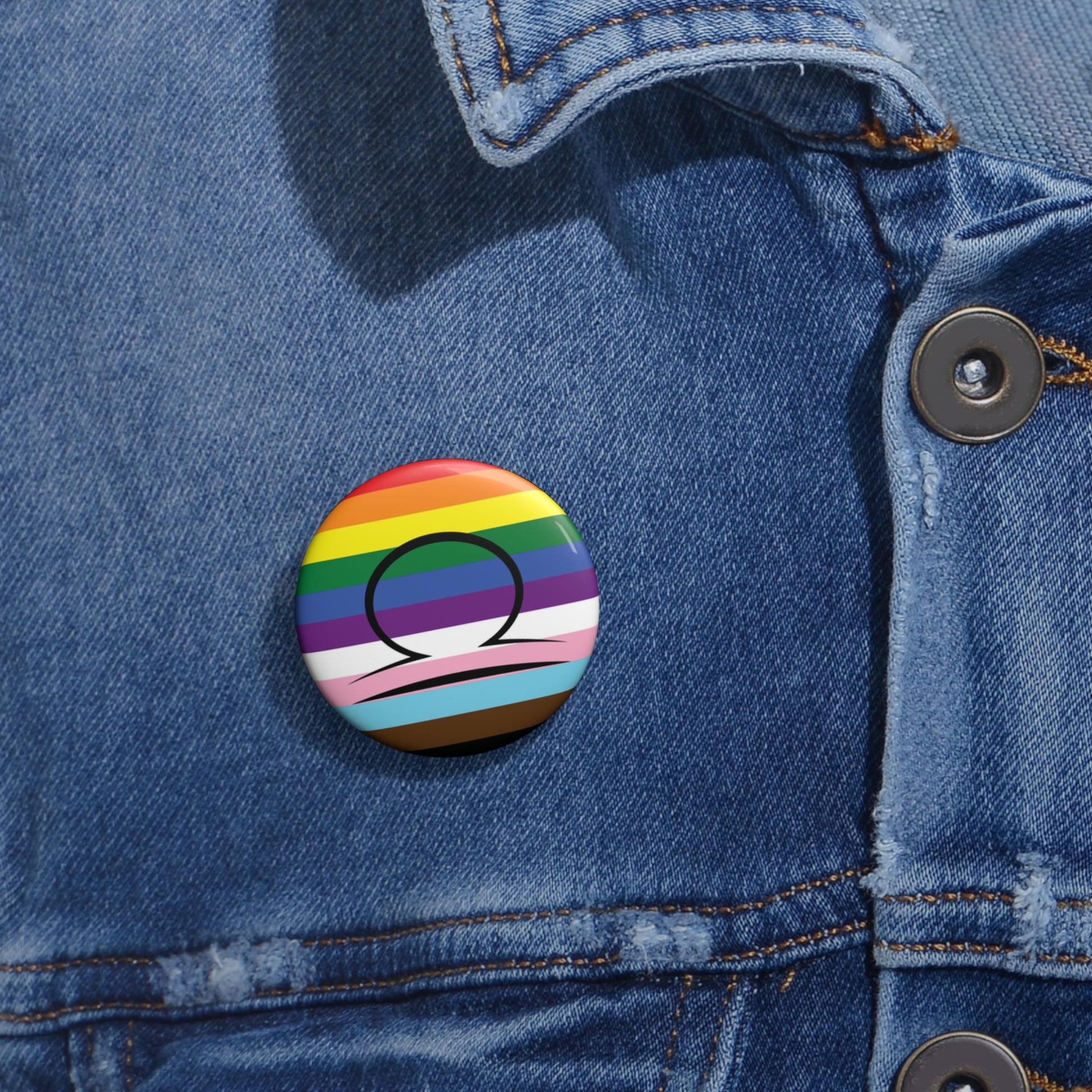 Close-up of a white circular pride button featuring the Libra zodiac glyph centered over a Progress Pride flag background in rainbow stripes with black, brown, light blue, pink, and white elements, attached to a blue denim jacket at the right chest with the button clearly visible.