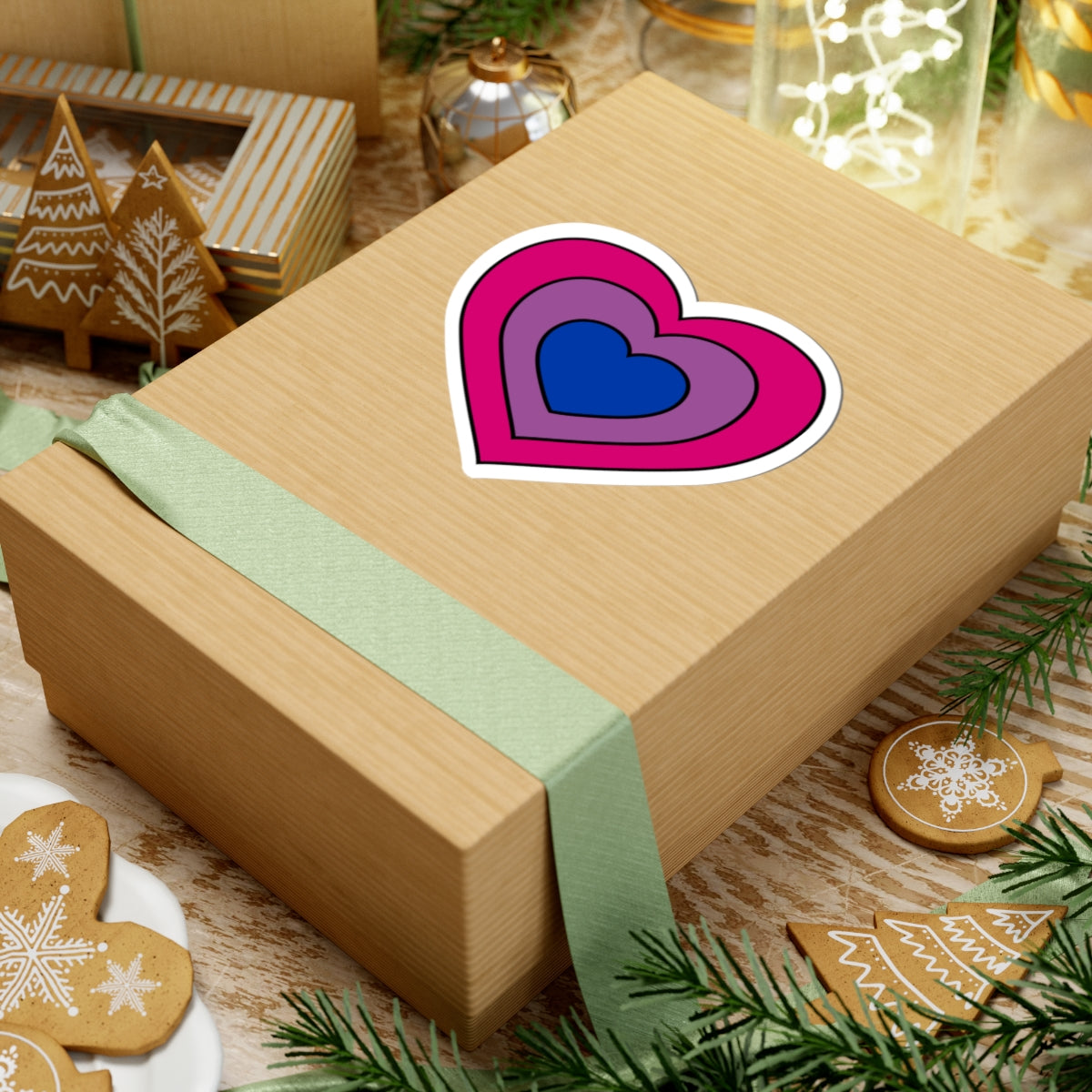 Gift box with a green ribbon on a festive background with cookies and Christmas decorations. Heart-shaped Bisexual pride sticker centered on the box.
