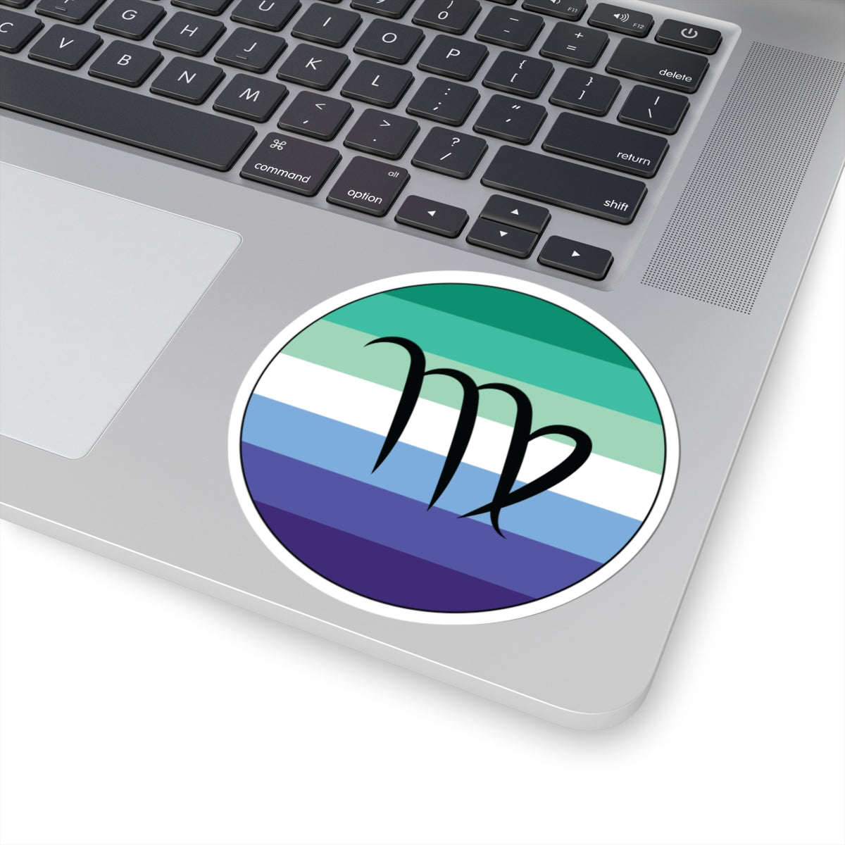 Close-up of the bottom right corner of a silver laptop featuring a 4 inch by 4 inch circular zodiac sticker with a centered Virgo glyph on a Gay Men pride flag style background applied to the surface.