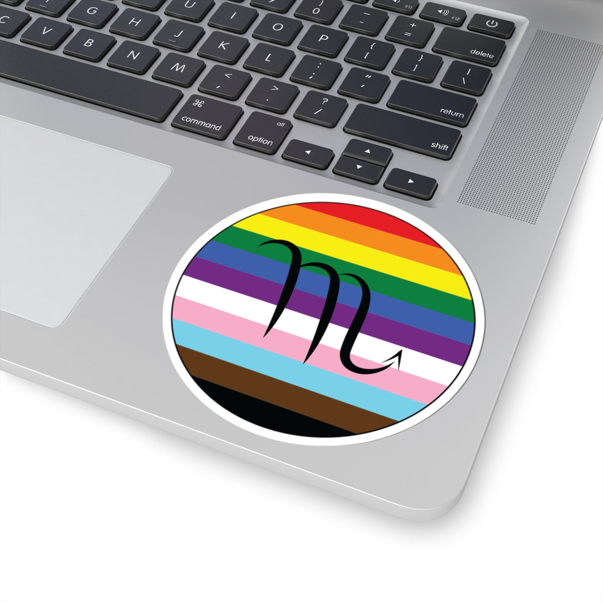 Close-up of the bottom right corner of a silver laptop featuring a 4 inch by 4 inch circular zodiac sticker with a centered Scorpio glyph on a progress pride flag style background applied to the surface.