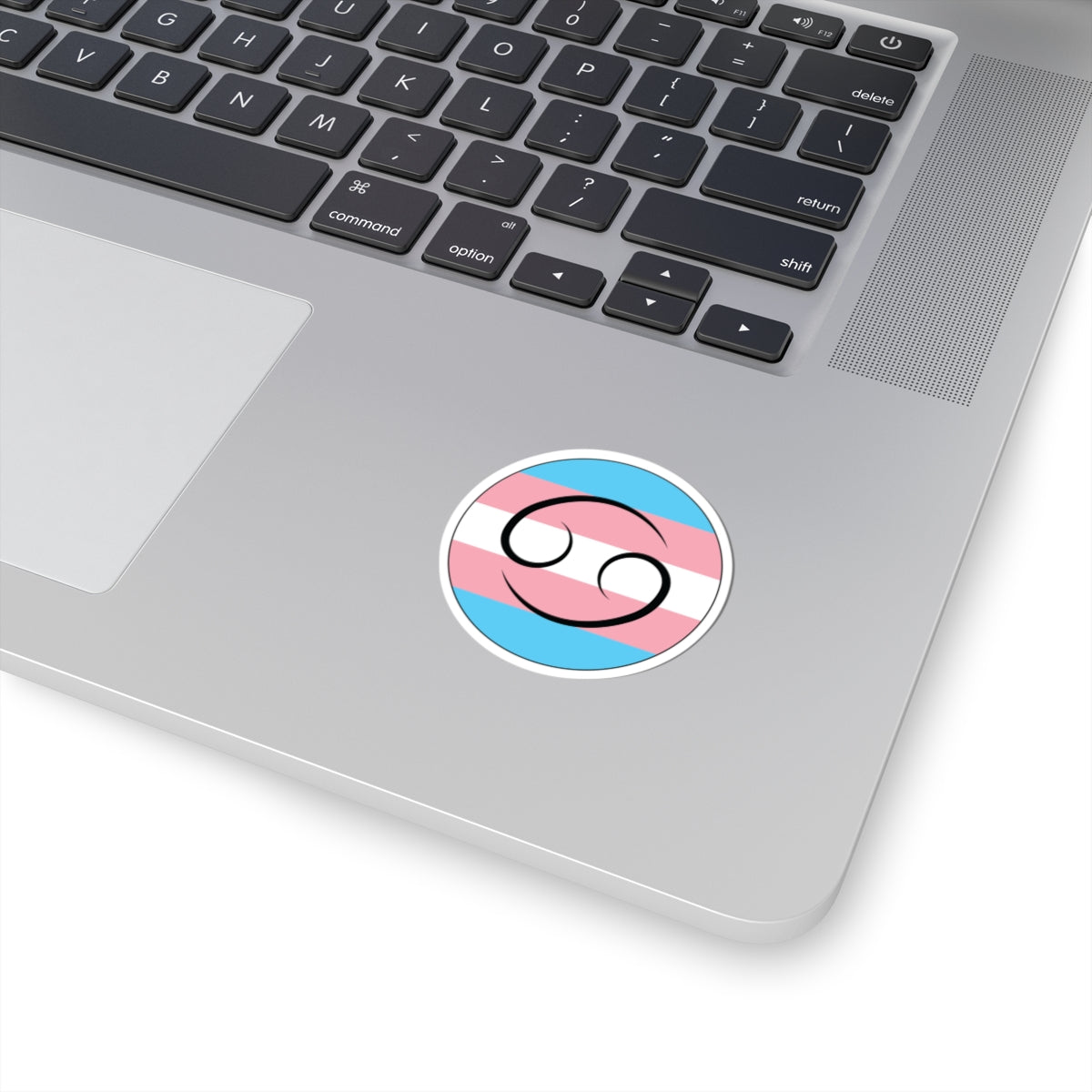 Close-up of the bottom right corner of a silver laptop featuring a 2 inch by 2 inch circular zodiac sticker with a centered Cancer glyph on a Transgender pride flag style background applied to the surface.